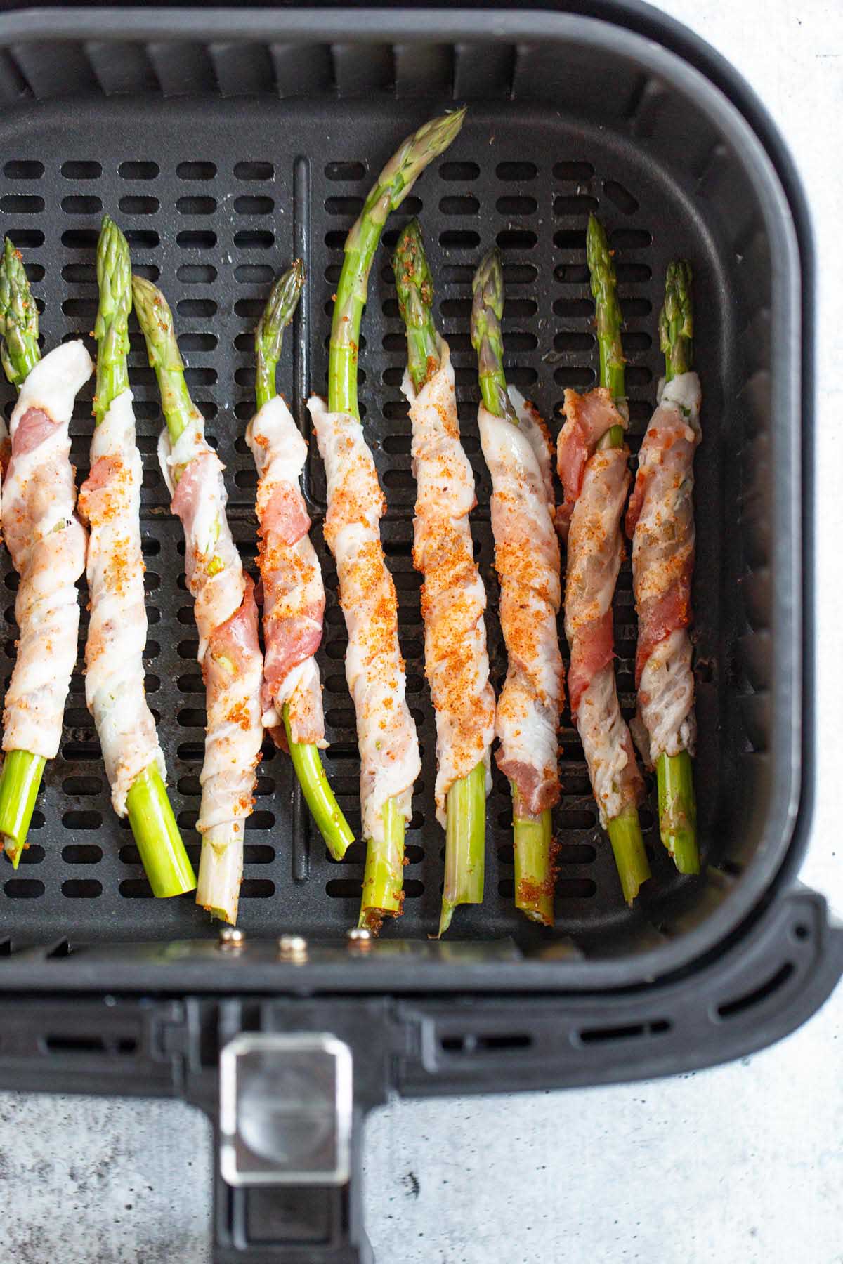 Uncooked Bacon wrapped asparagus in air fryer basket.